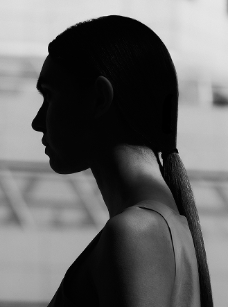 Black and white profile of a person with a sleek ponytail, face and front in deep shadow.