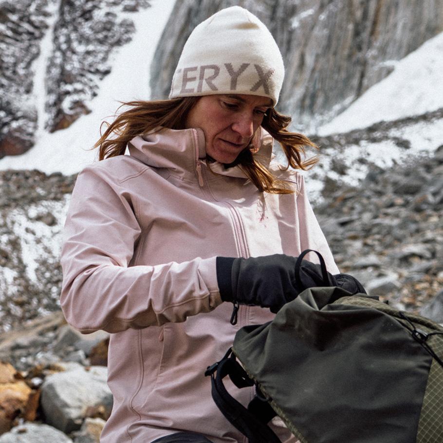 A woman in a pink jacket and Arc'teryx beanie packing a green backpack in a snowy mountain environment.