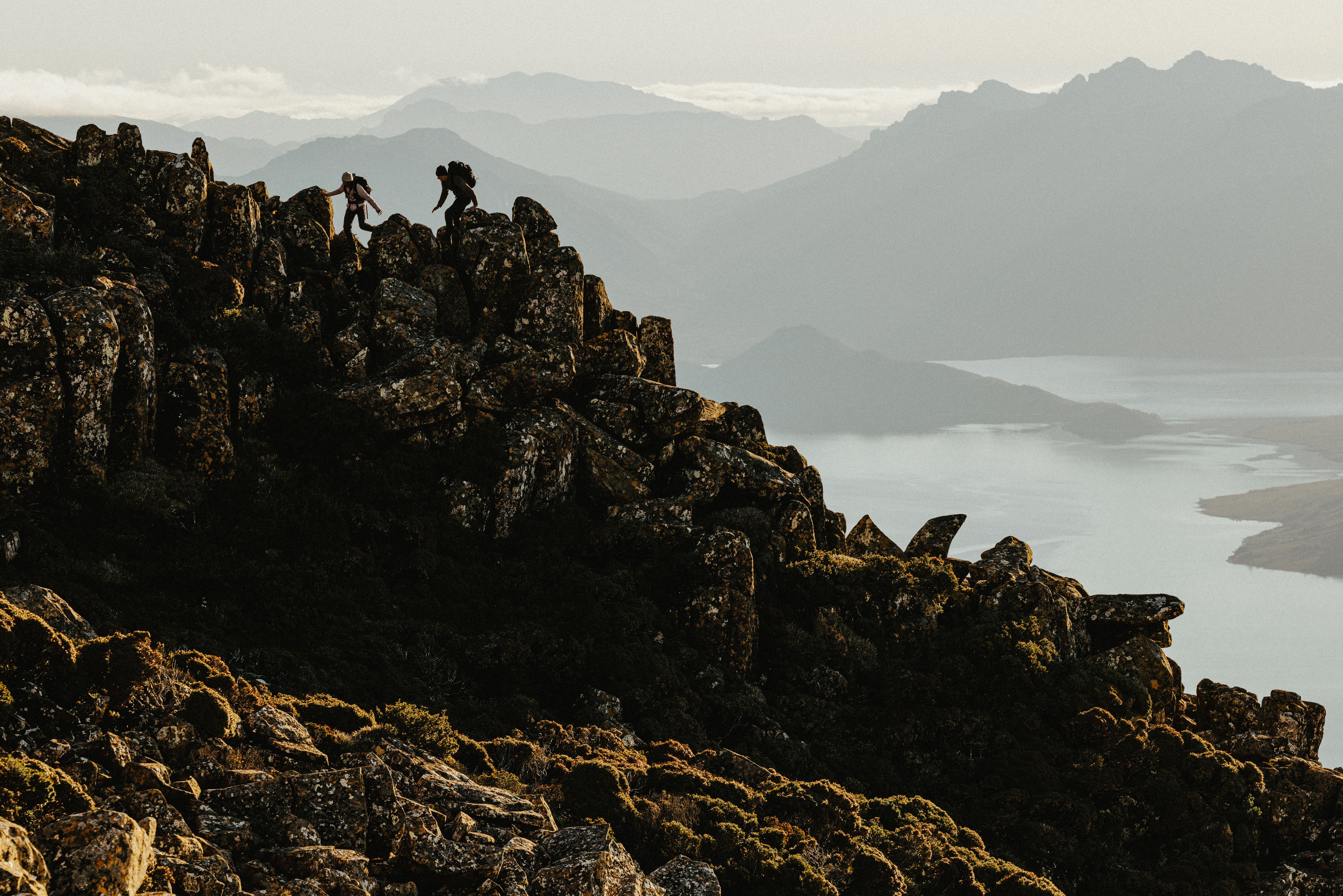 Two hikers on a rocky mountain ridge overlooking a scenic lake and distant peaks.