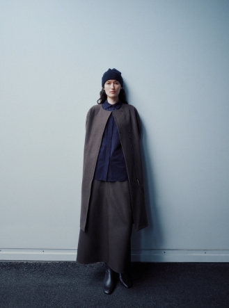 A woman in a dark beanie, navy shirt, long dark gray cape, and skirt stands against a light blue wall.