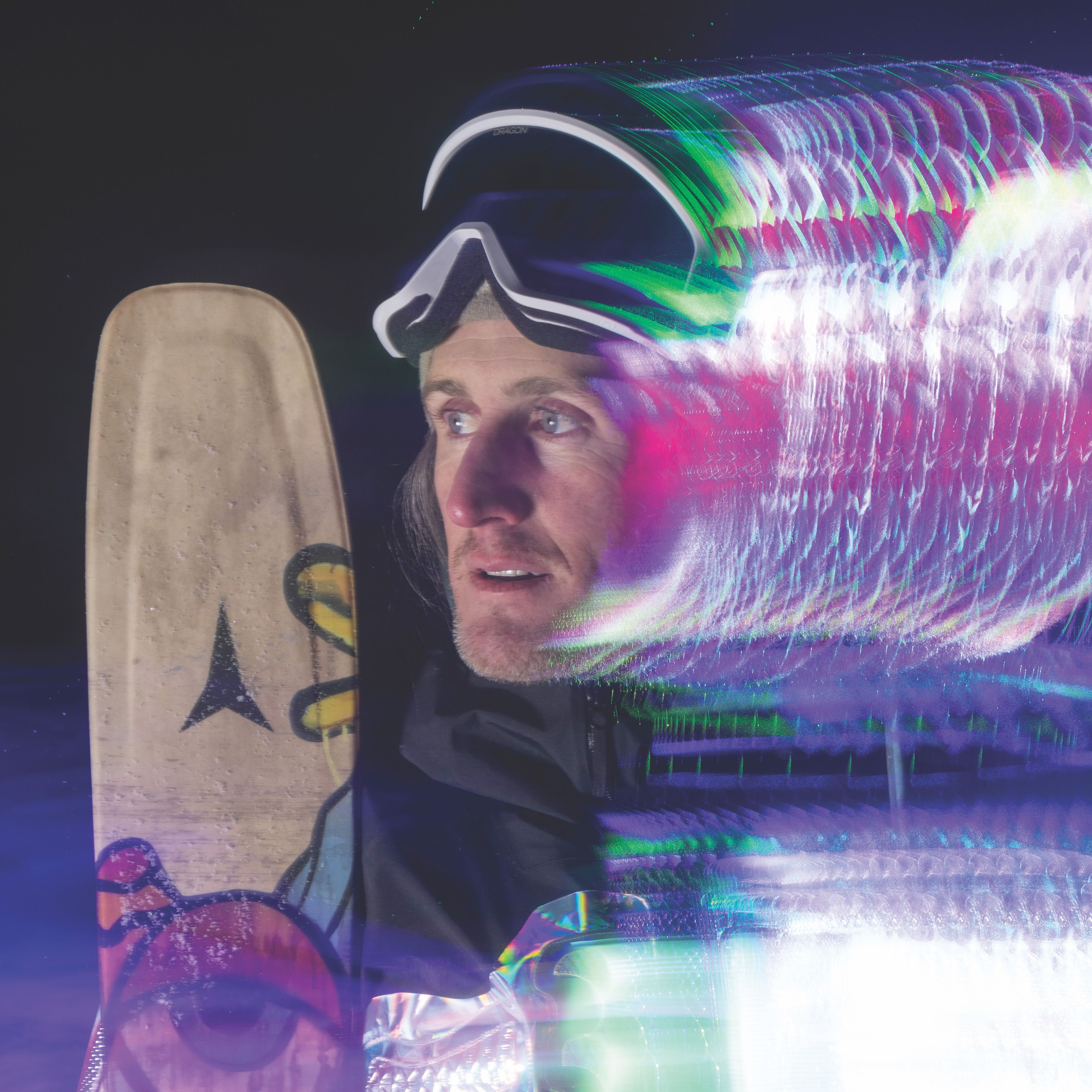 Chris Benchetler in ski goggles looking left, with a snowboard and a colorful light trail effect on the right.