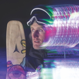 Chris Benchetler in ski goggles looking left, with a snowboard and a colorful light trail effect on the right.