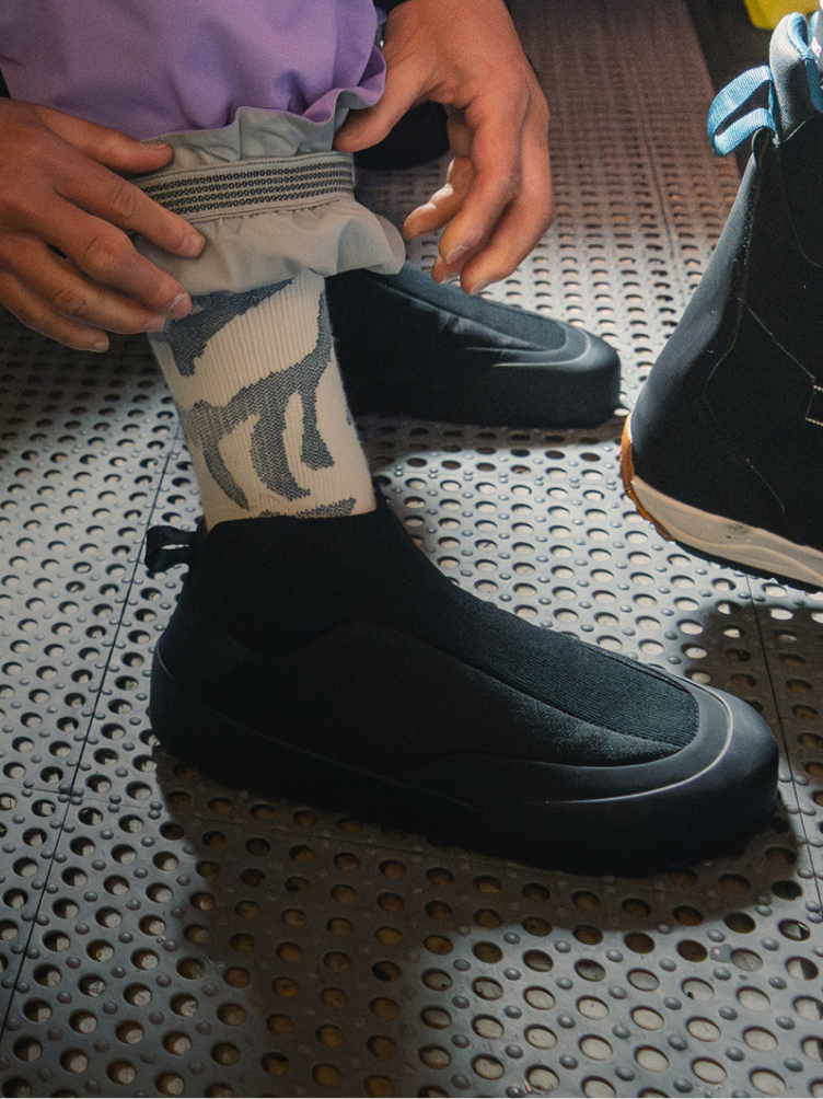 A person adjusts their pant cuff above a patterned sock and a black slip-on shoe on a perforated floor.