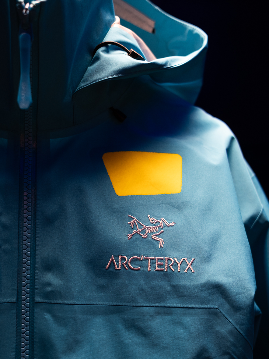 A close-up of a teal Arc'teryx jacket with a pink embroidered logo and a yellow chest patch.