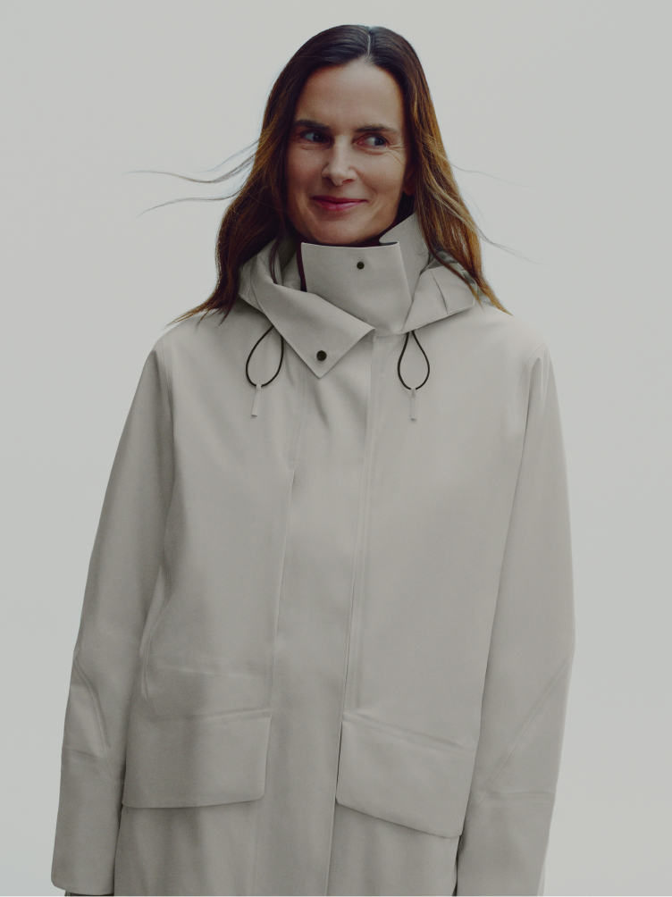 A smiling woman with brown hair wears an off-white hooded jacket.