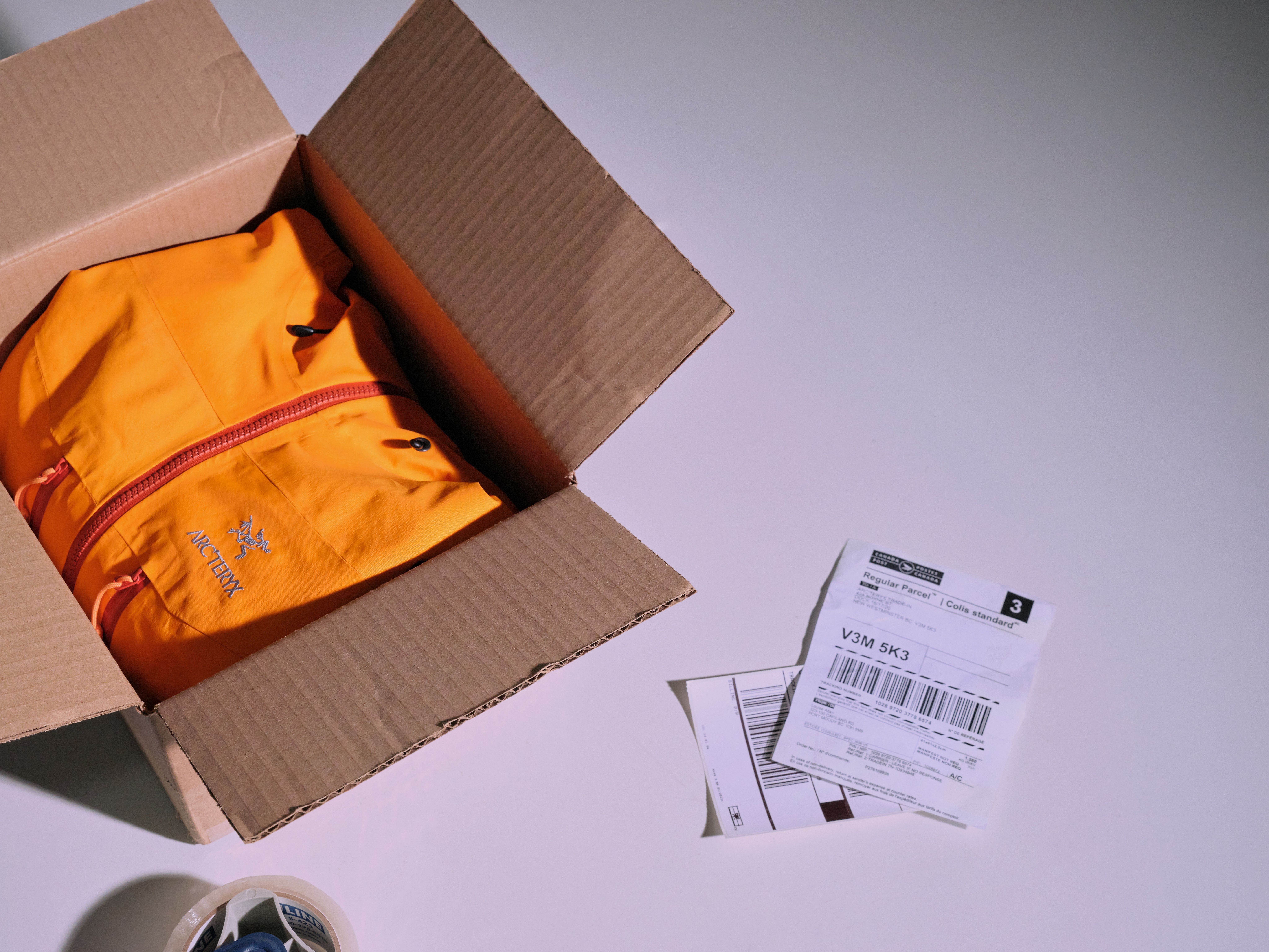 An orange Arc'teryx jacket packed in an open cardboard box with shipping labels nearby.