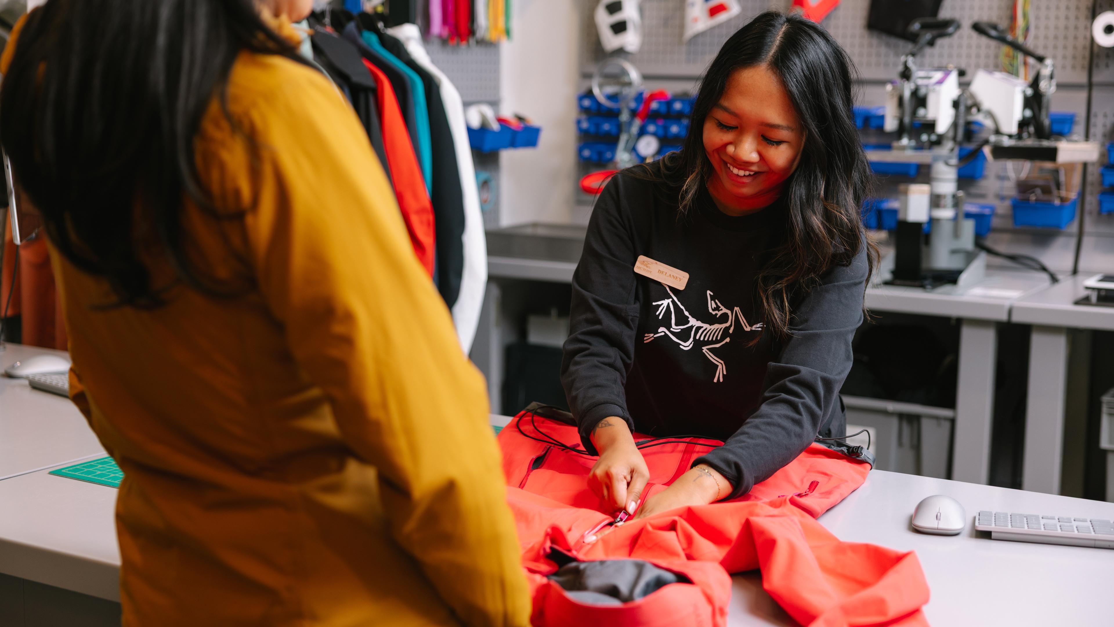 A smiling Arc'teryx employee repairs a red jacket for a customer.