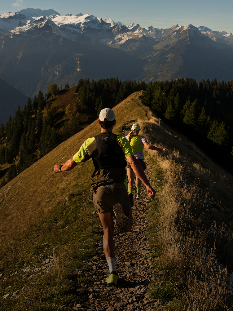 Two people trail running along a narrow mountain ridge with snow-capped peaks in the background.