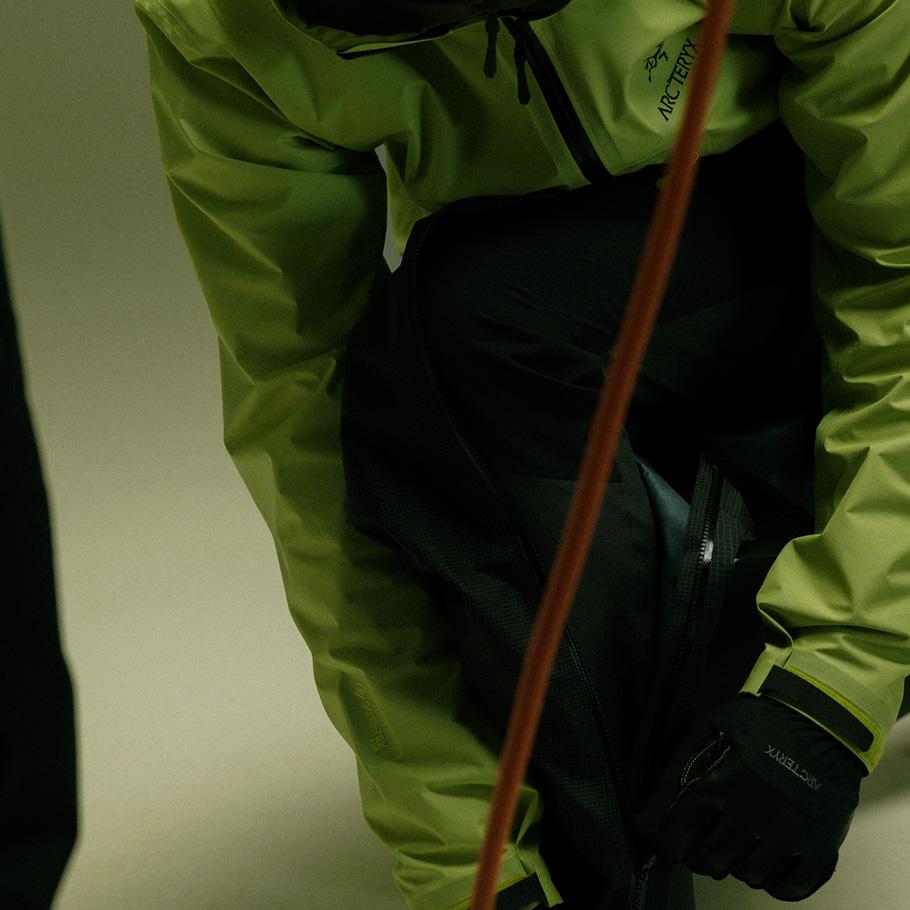 A person in a lime green Arc'teryx jacket and dark pants adjusts a zipper, an orange rope crosses the frame.