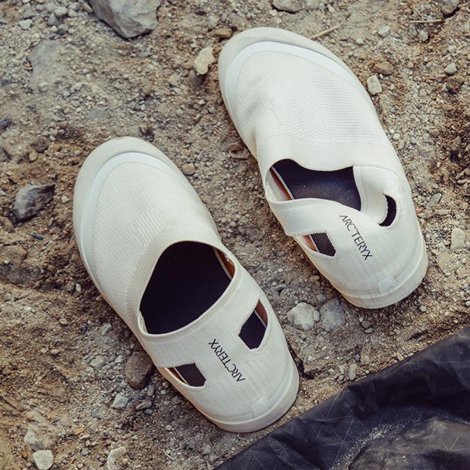 A pair of cream-colored Arc'teryx slip-on shoes on a gravel surface.