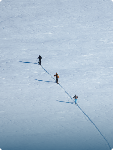 Three people ski touring in single file across a vast snowfield, casting long shadows.