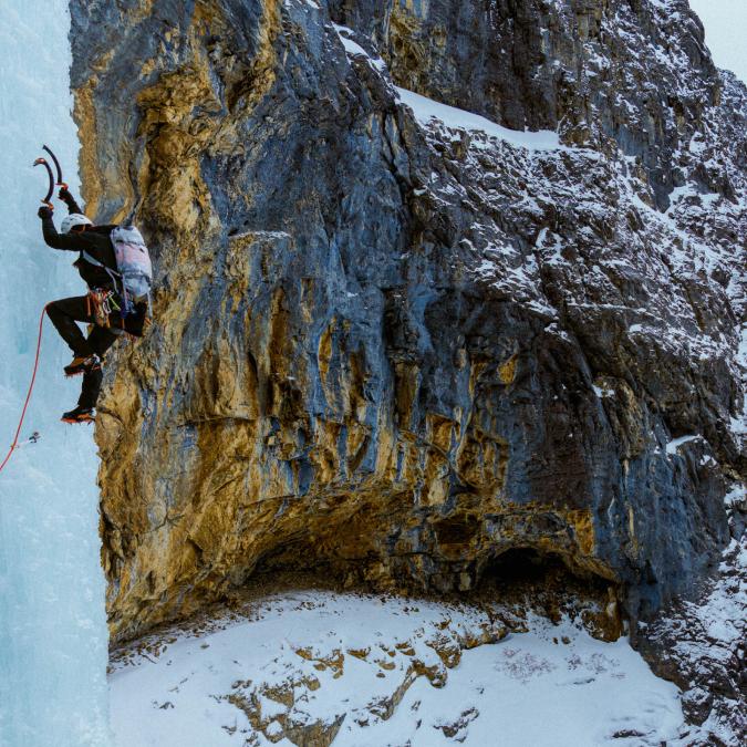 An ice climber ascends a tall ice formation beside a snowy rock face.
