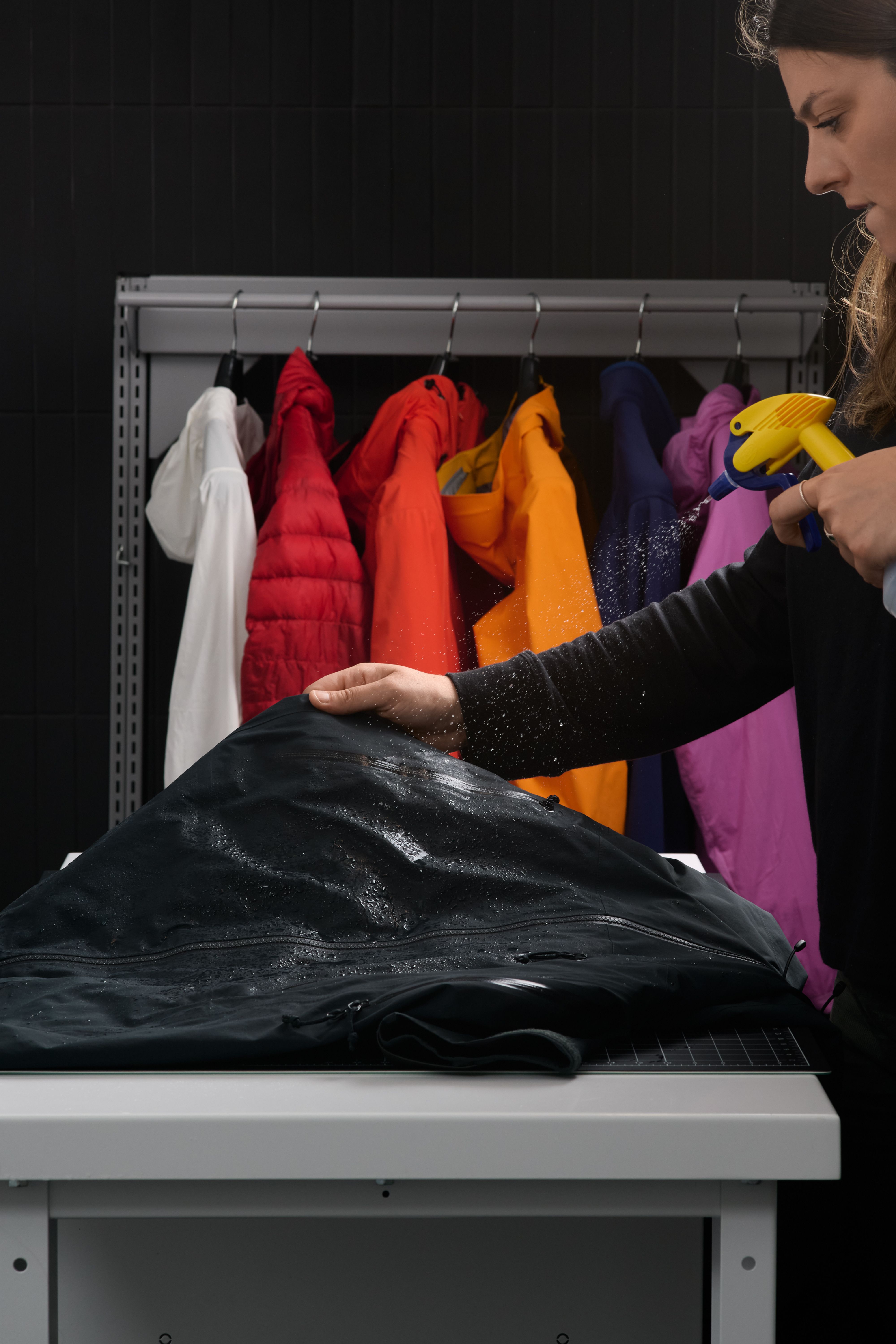 A woman sprays water onto a black waterproof jacket on a table, with a rack of colorful jackets in the background.