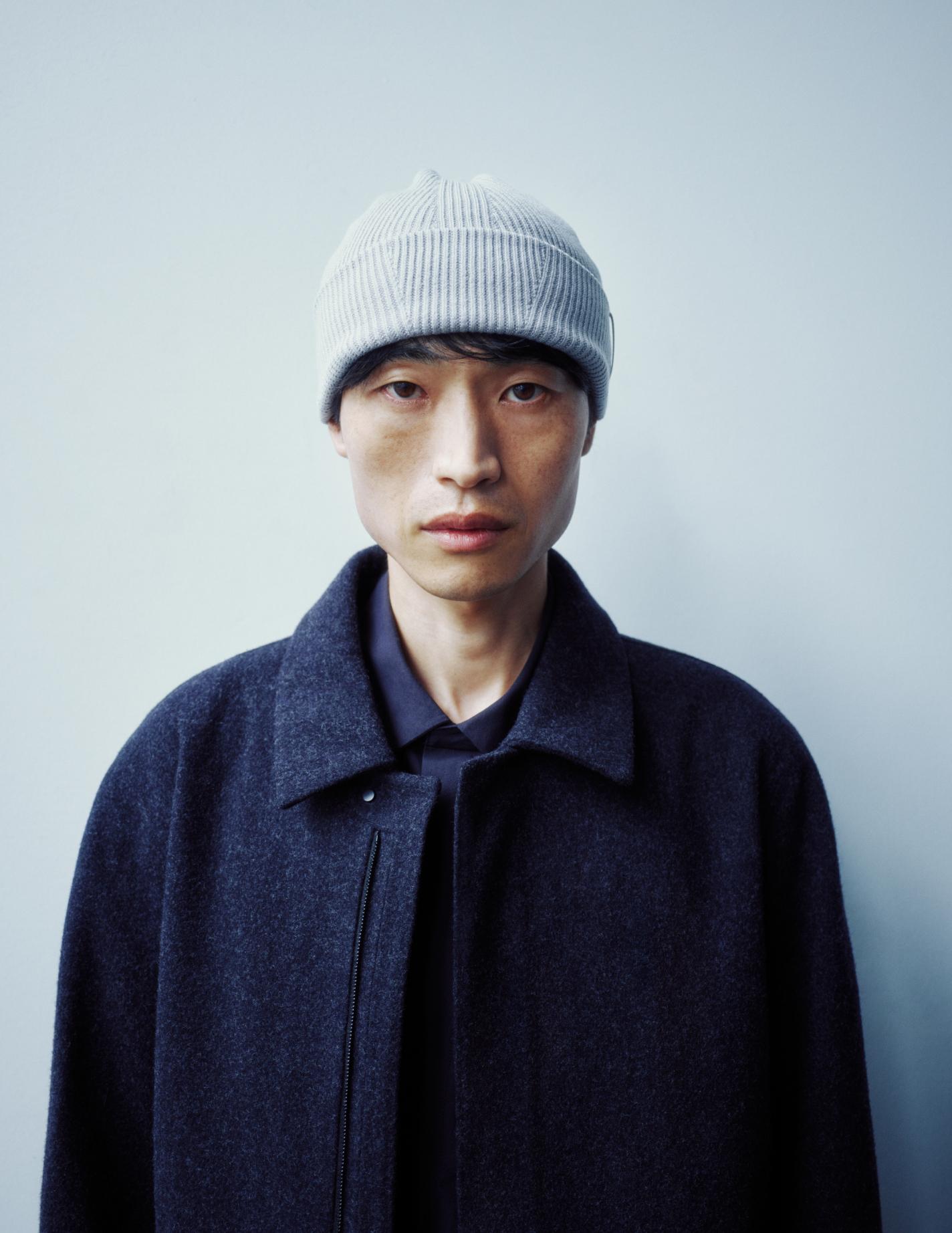 A man in a light gray beanie, navy shirt, and dark blue wool coat looks directly at the viewer.