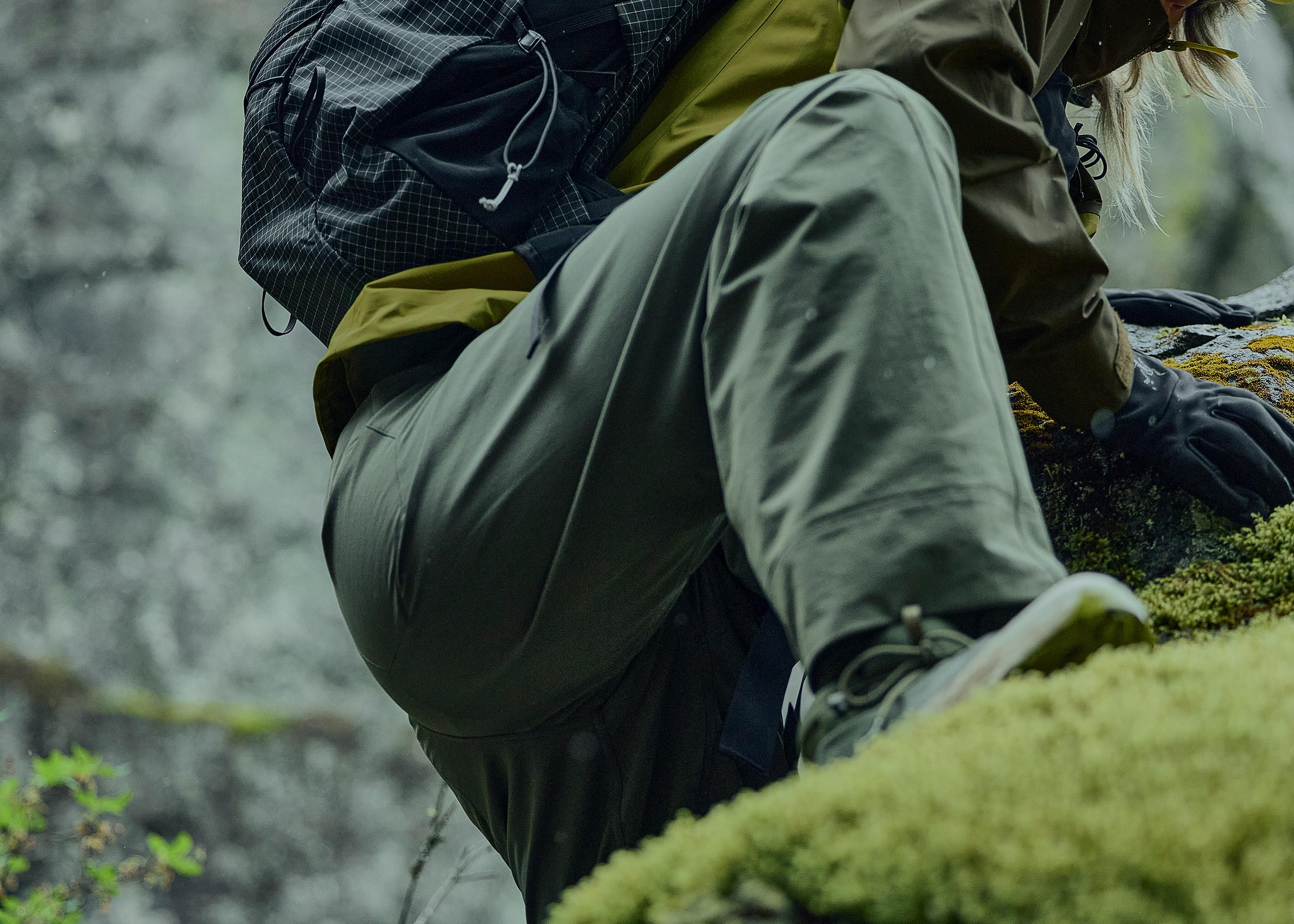 A hiker in olive green pants and a backpack climbing over a mossy rock.