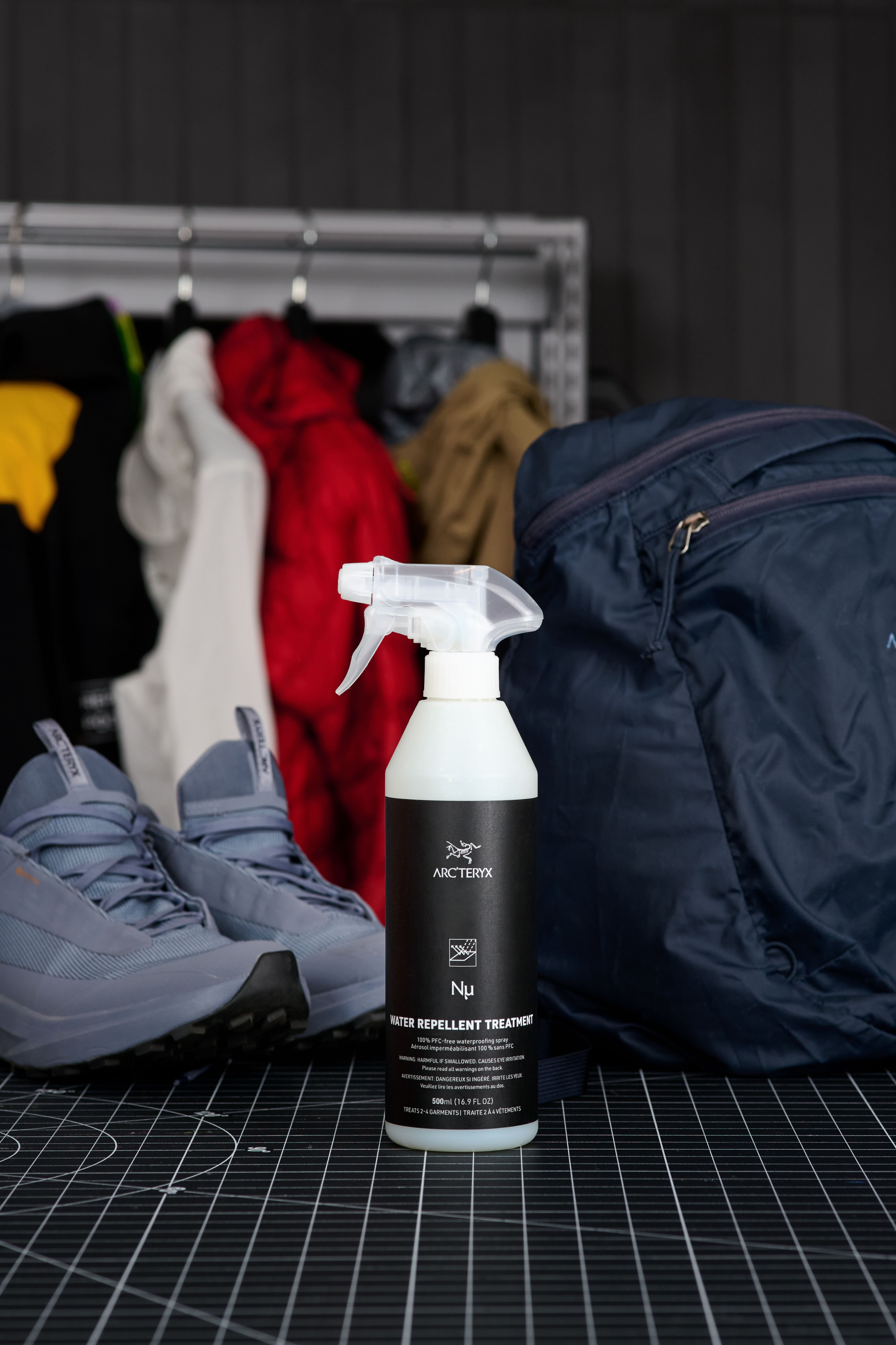 An Arc'teryx water repellent treatment spray bottle on a grid mat, flanked by grey sneakers and a blue backpack, with jackets in the blurred background.