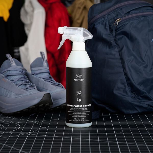 An Arc'teryx water repellent treatment spray bottle on a grid mat, flanked by grey sneakers and a blue backpack, with jackets in the blurred background.