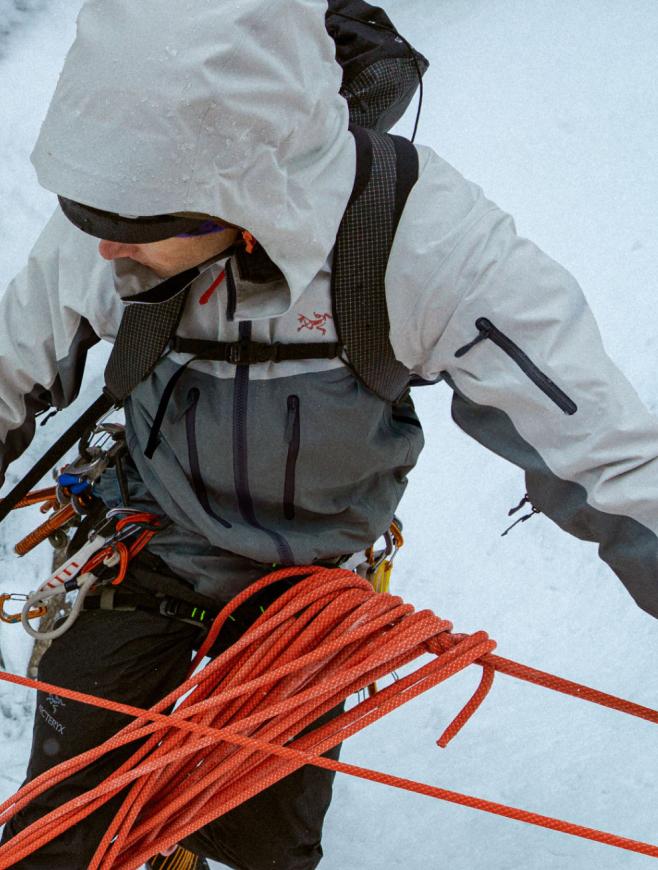Close-up of a climber in a grey hooded jacket, wearing a harness with gear, and holding a coiled orange rope.