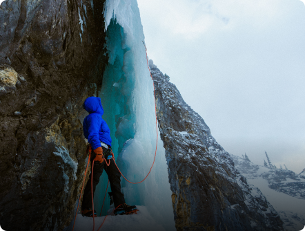 An ice climber in a blue jacket stands at the base of a towering blue ice formation, ready to ascend.