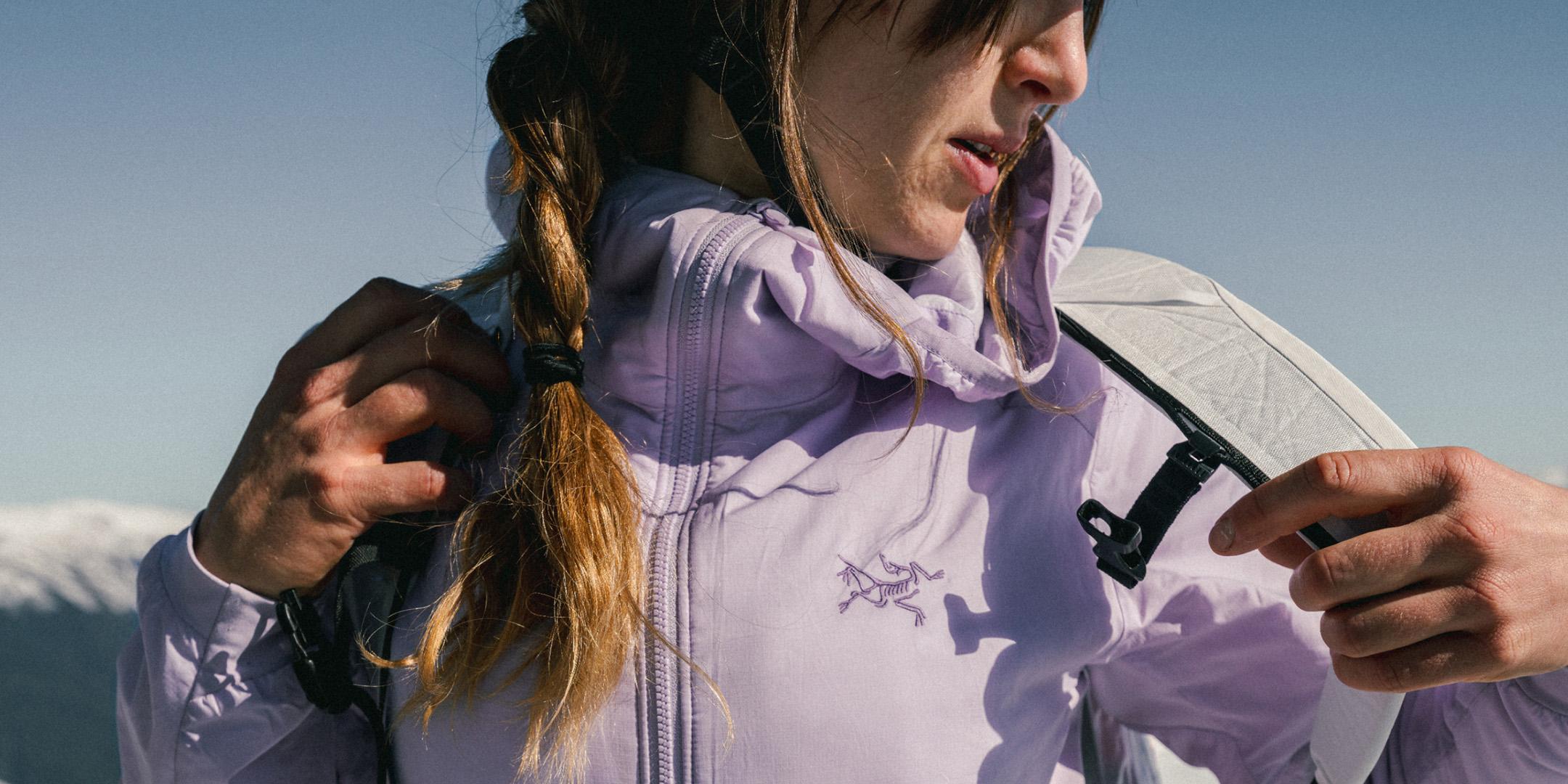 Woman in a light purple Arc'teryx jacket adjusting a strap, with mountains in the background.