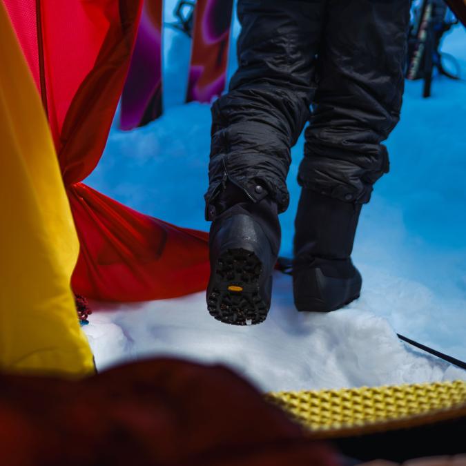 Person in black snow boots stepping out of a red and yellow tent into fresh snow, showing a Vibram sole.