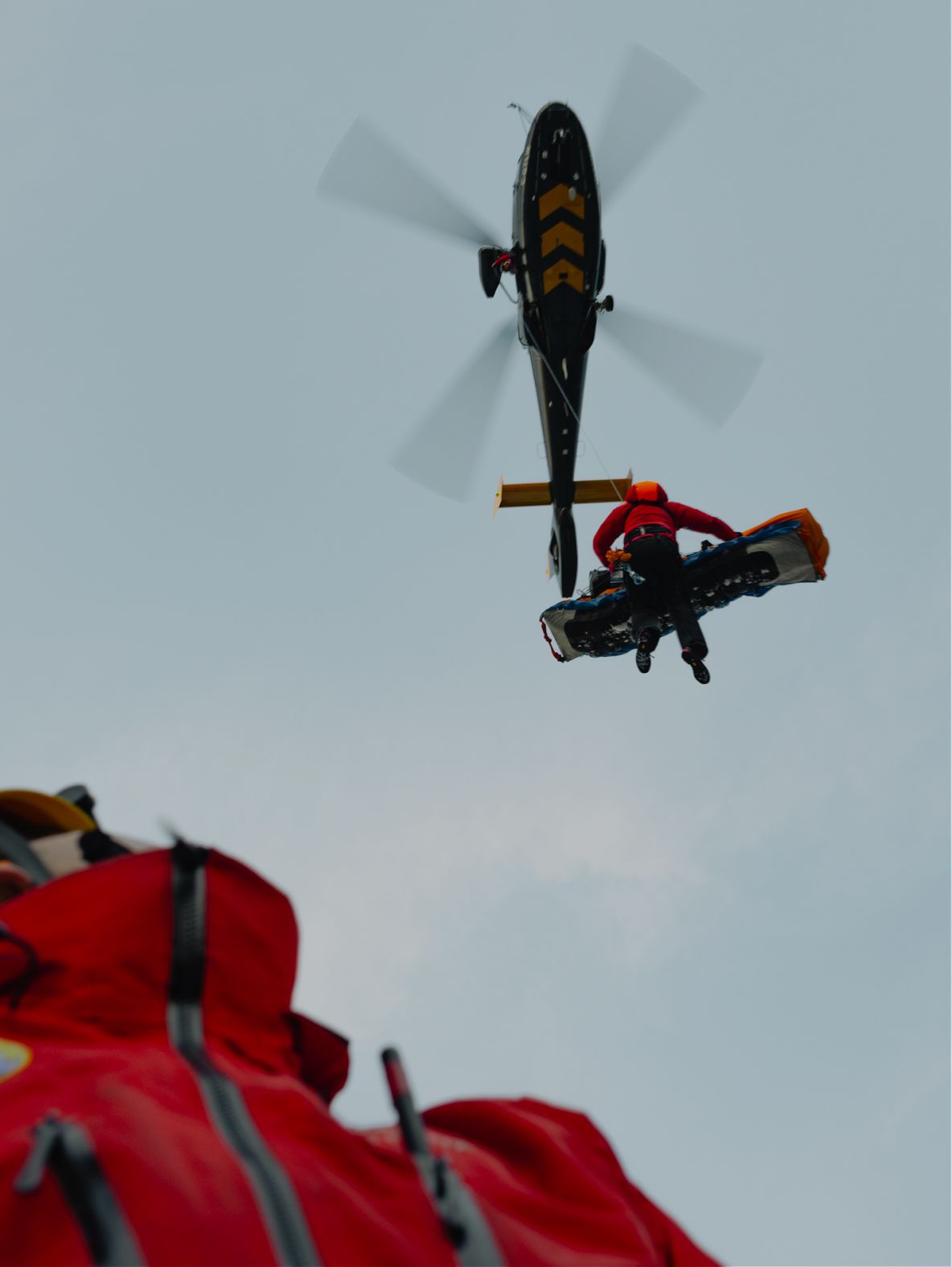 A rescuer suspended from a helicopter carries a stretcher under an overcast sky, with a red jacket in the foreground.