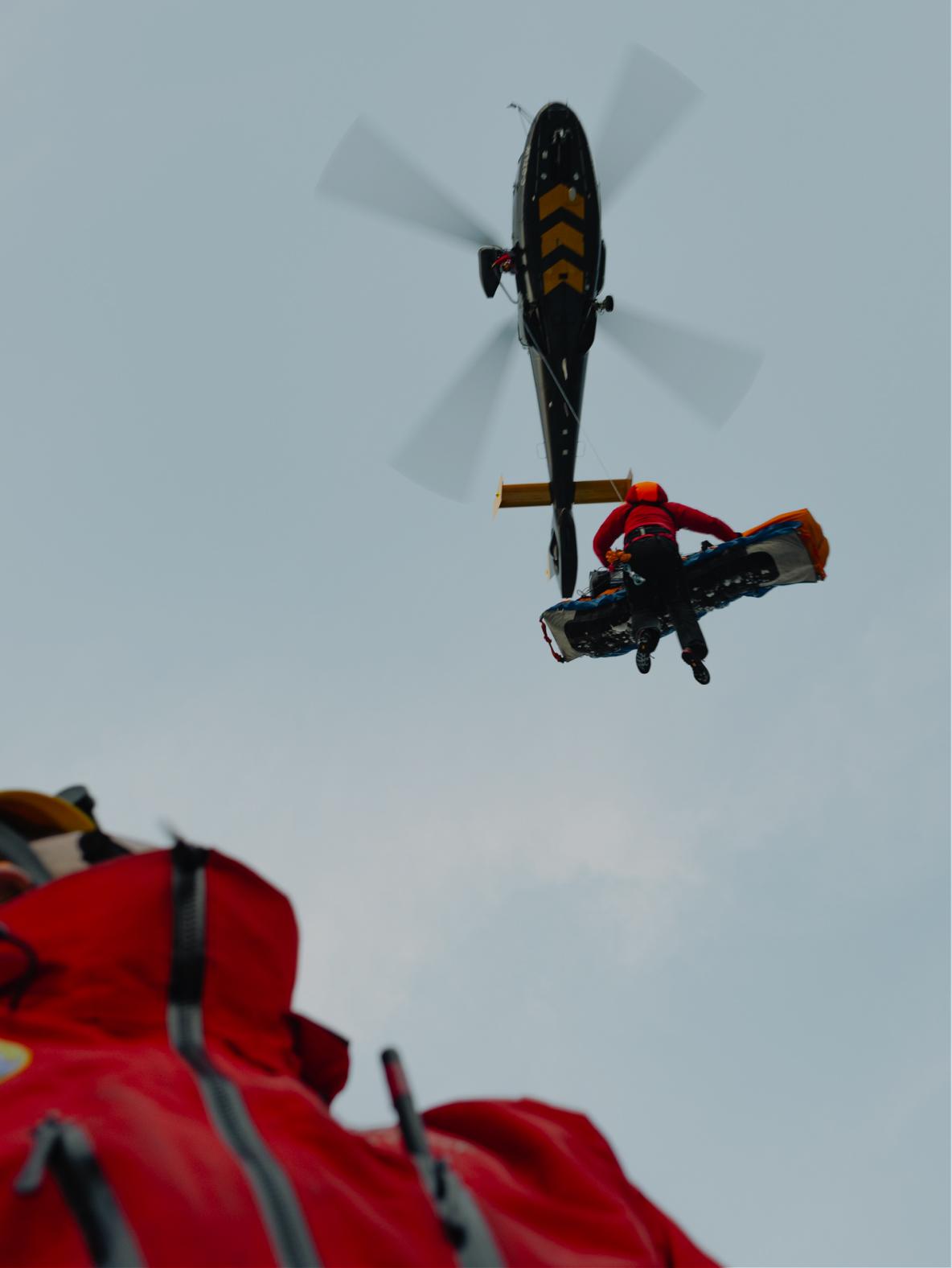 A rescuer suspended from a helicopter carries a stretcher under an overcast sky, with a red jacket in the foreground.