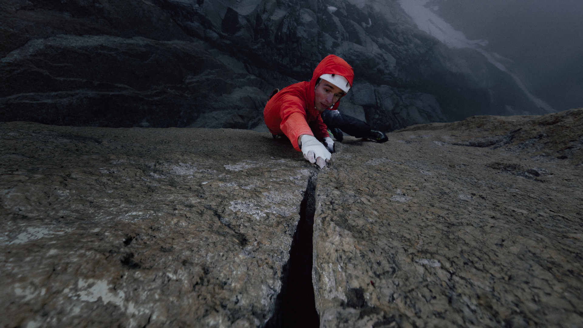 A climber in a red jacket and white gloves ascends a vertical rock crack, with a vast mountain range in the distance.