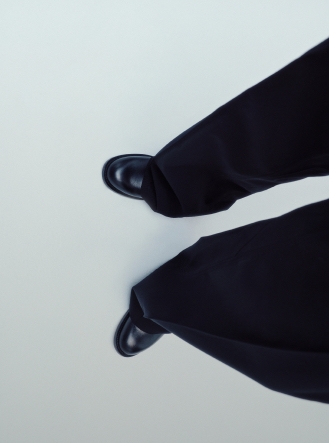 Overhead view of a person's legs in dark pants and black shoes on a light floor.