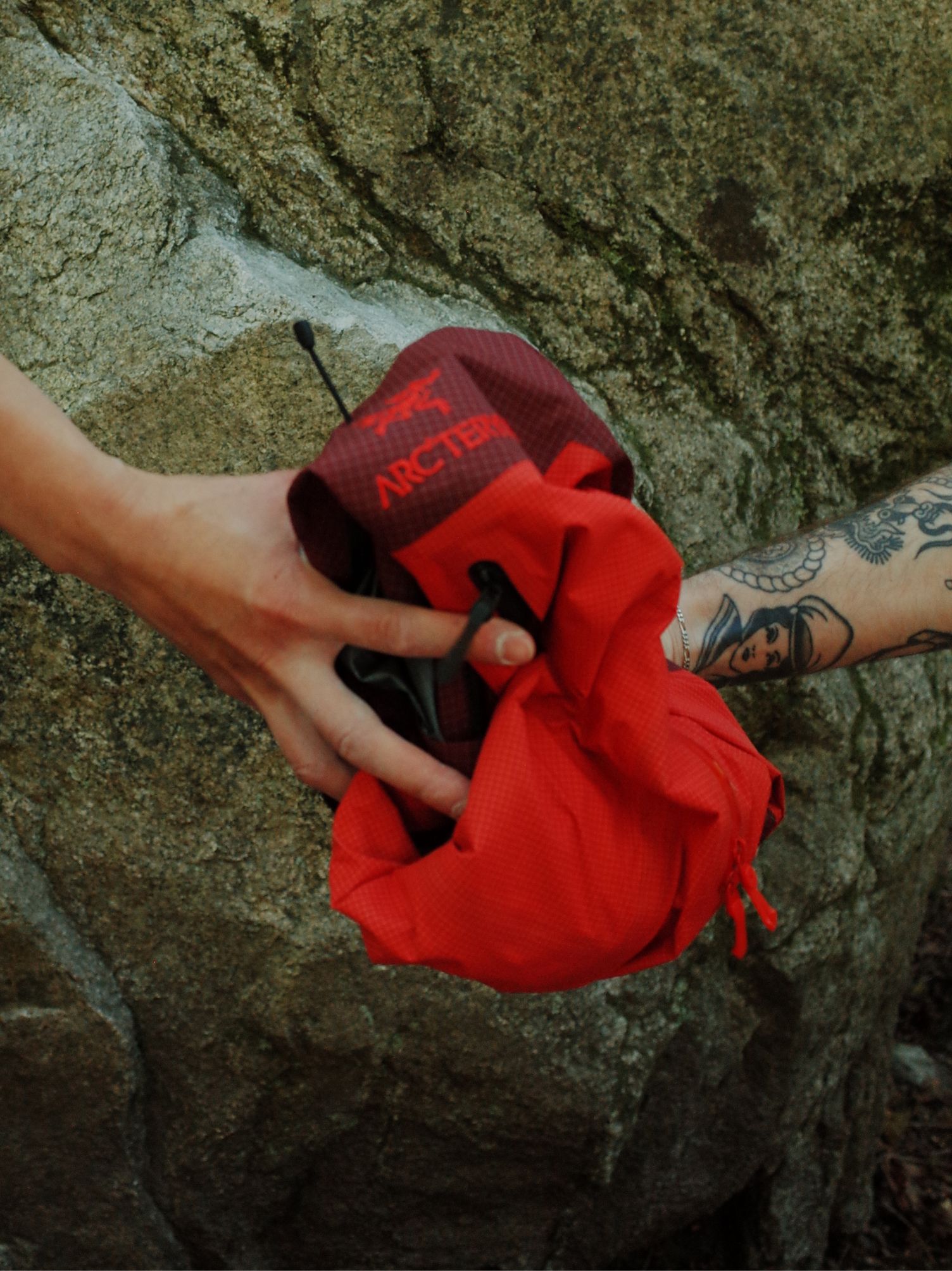 Two hands, one tattooed, exchanging a red Arc'teryx bag against a rock.
