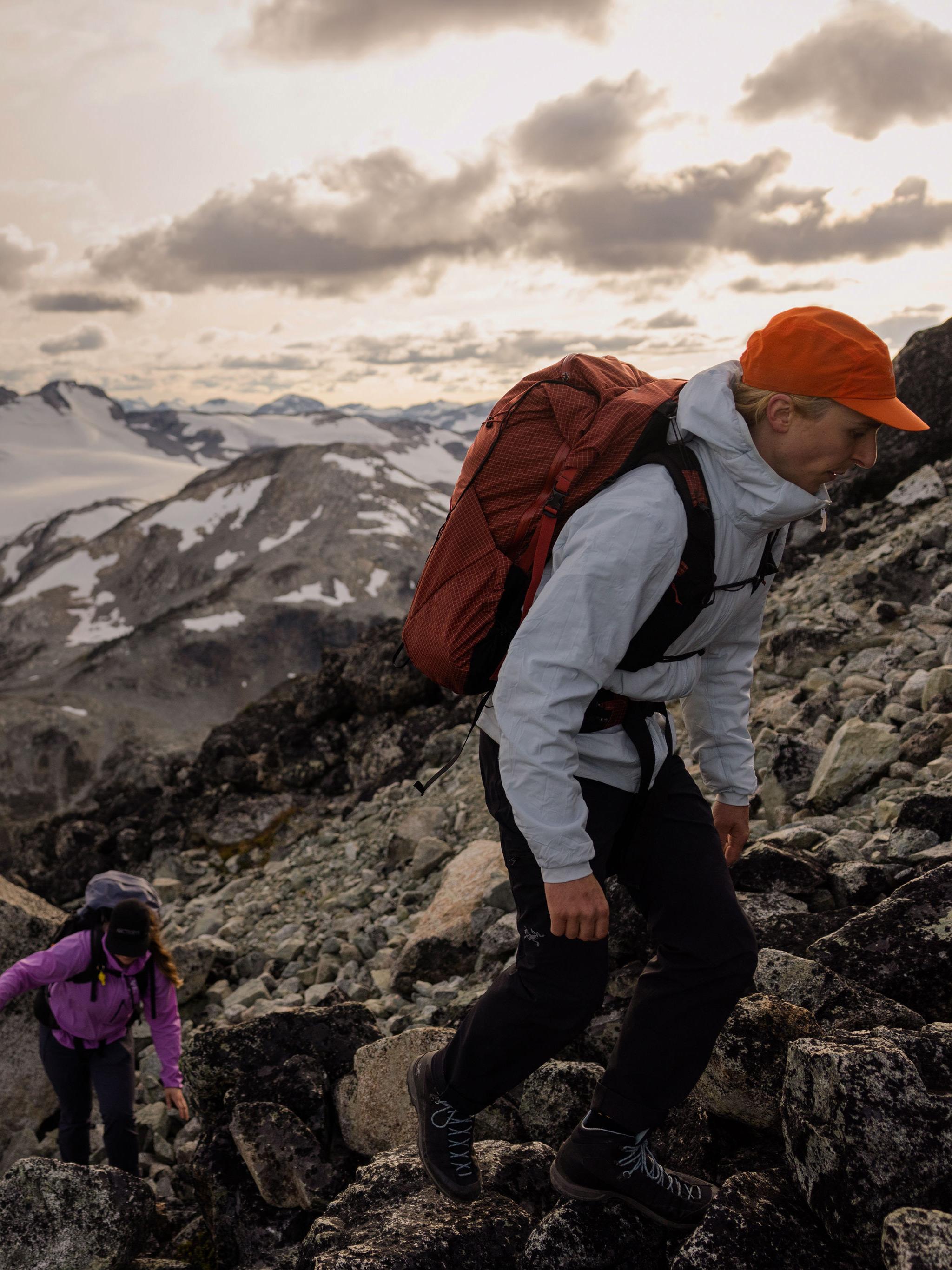 Two hikers ascend a rocky slope toward snow-capped mountains under a cloudy sky.