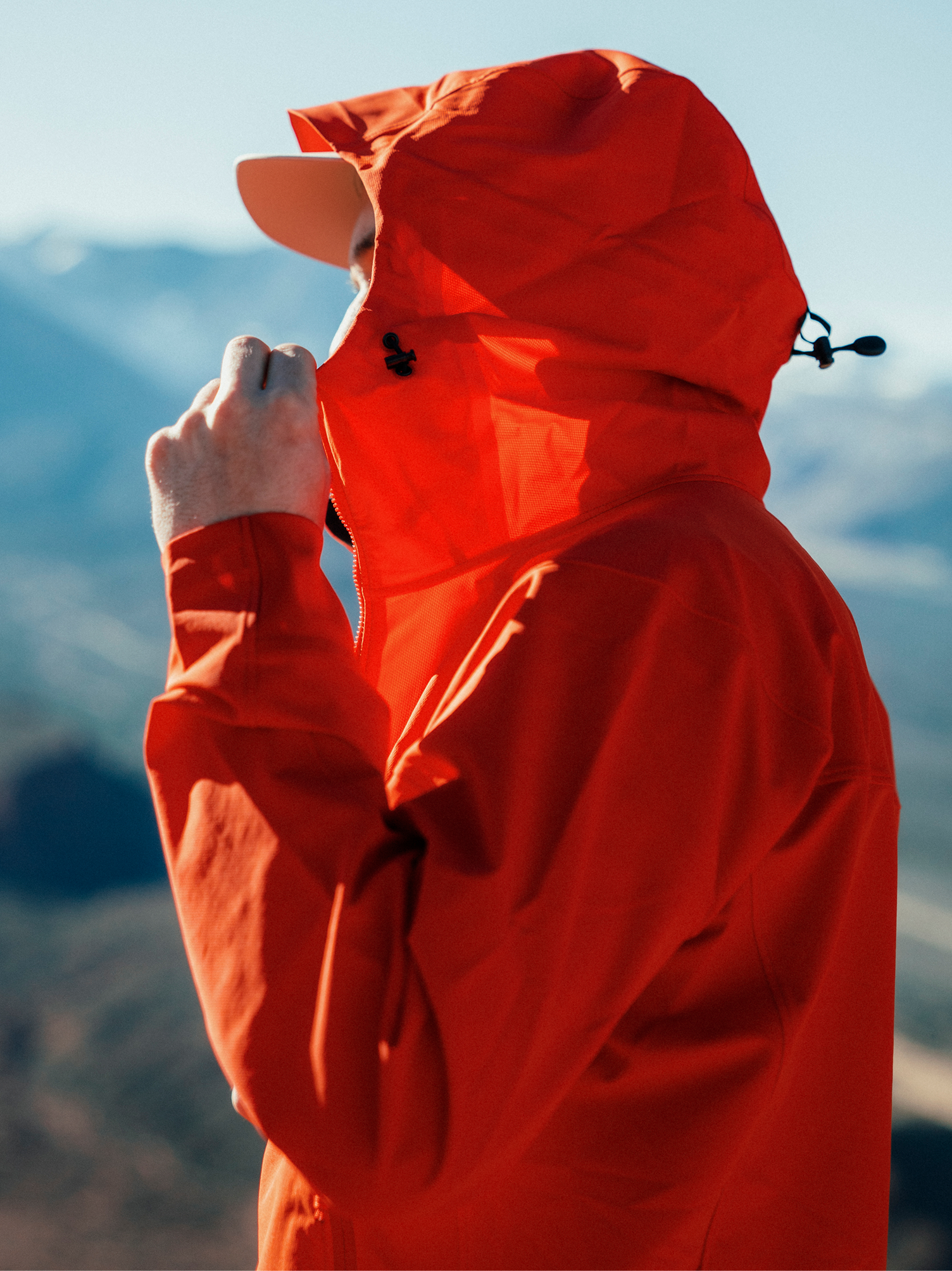 A person in a bright orange hooded jacket adjusts their collar outdoors.