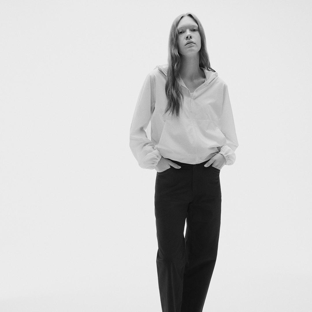 Black and white photo of a woman in a light hooded jacket and dark pants, standing with hands in pockets on a white background.