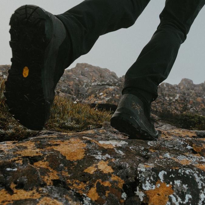 A person's hiking boots with Vibram and Arc'teryx logos stepping on rocks covered in yellow and white lichen.