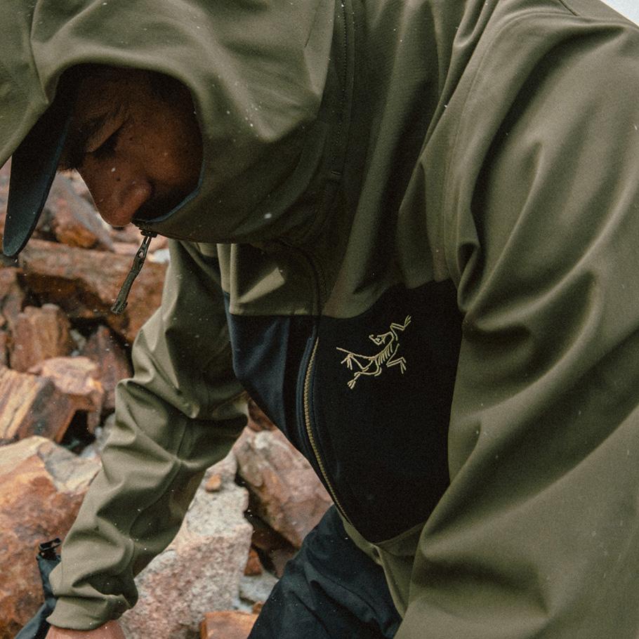 A person in an olive green Arc'teryx jacket with its hood up, looking down in a rocky outdoor environment.