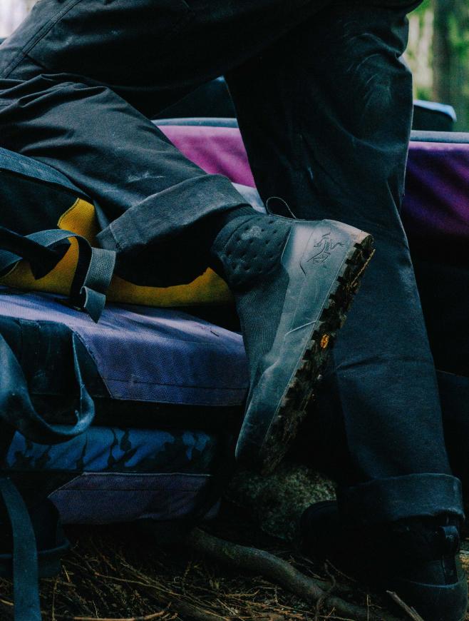 A person's legs in dark pants, with a rugged Arc'teryx shoe and yellow climbing shoes resting on purple crash pads on a forest floor.