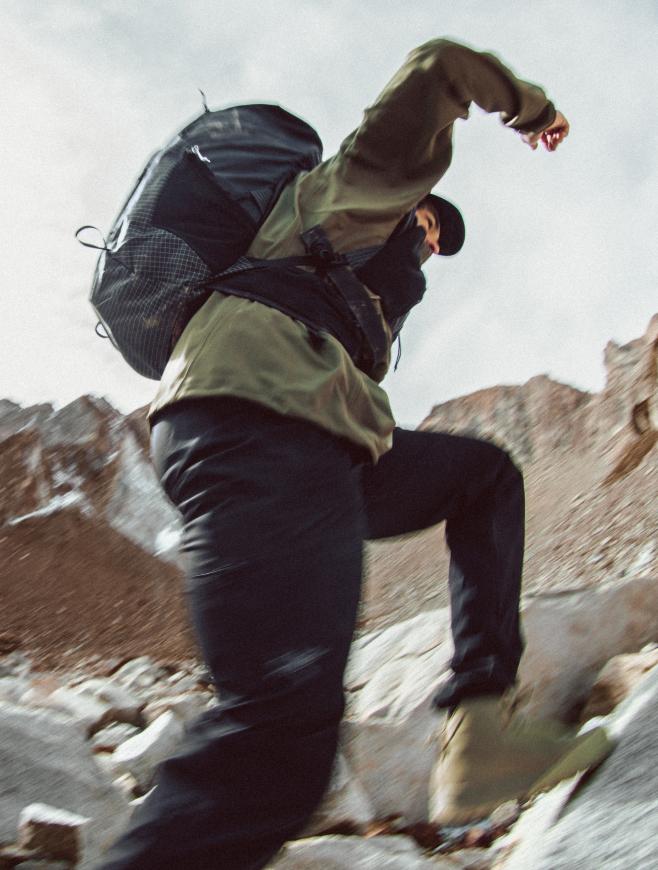 Hiker in a green jacket and black backpack quickly stepping up rocky terrain.