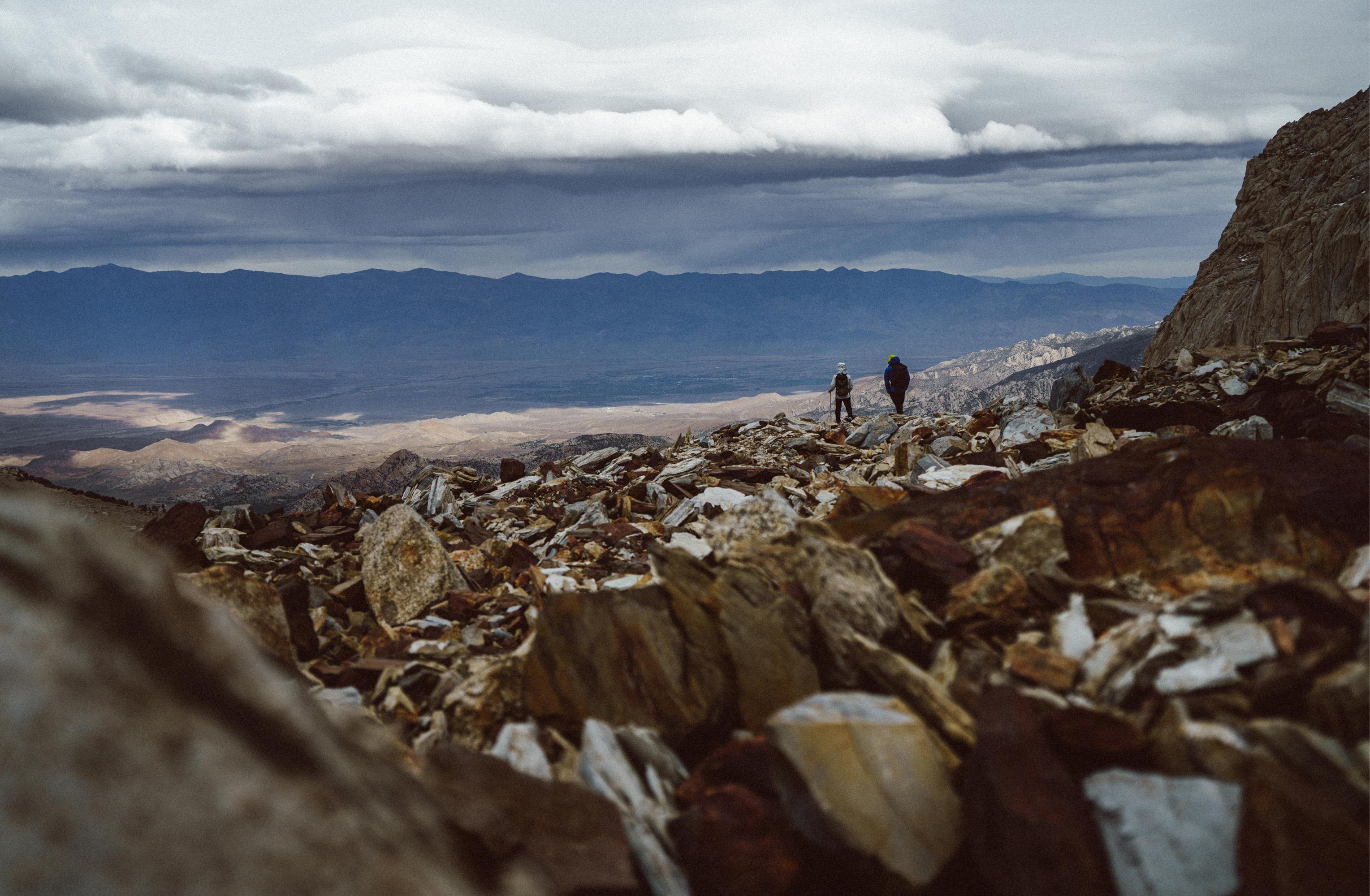 Two hikers on a rocky mountain slope overlooking a vast valley under a cloudy sky.