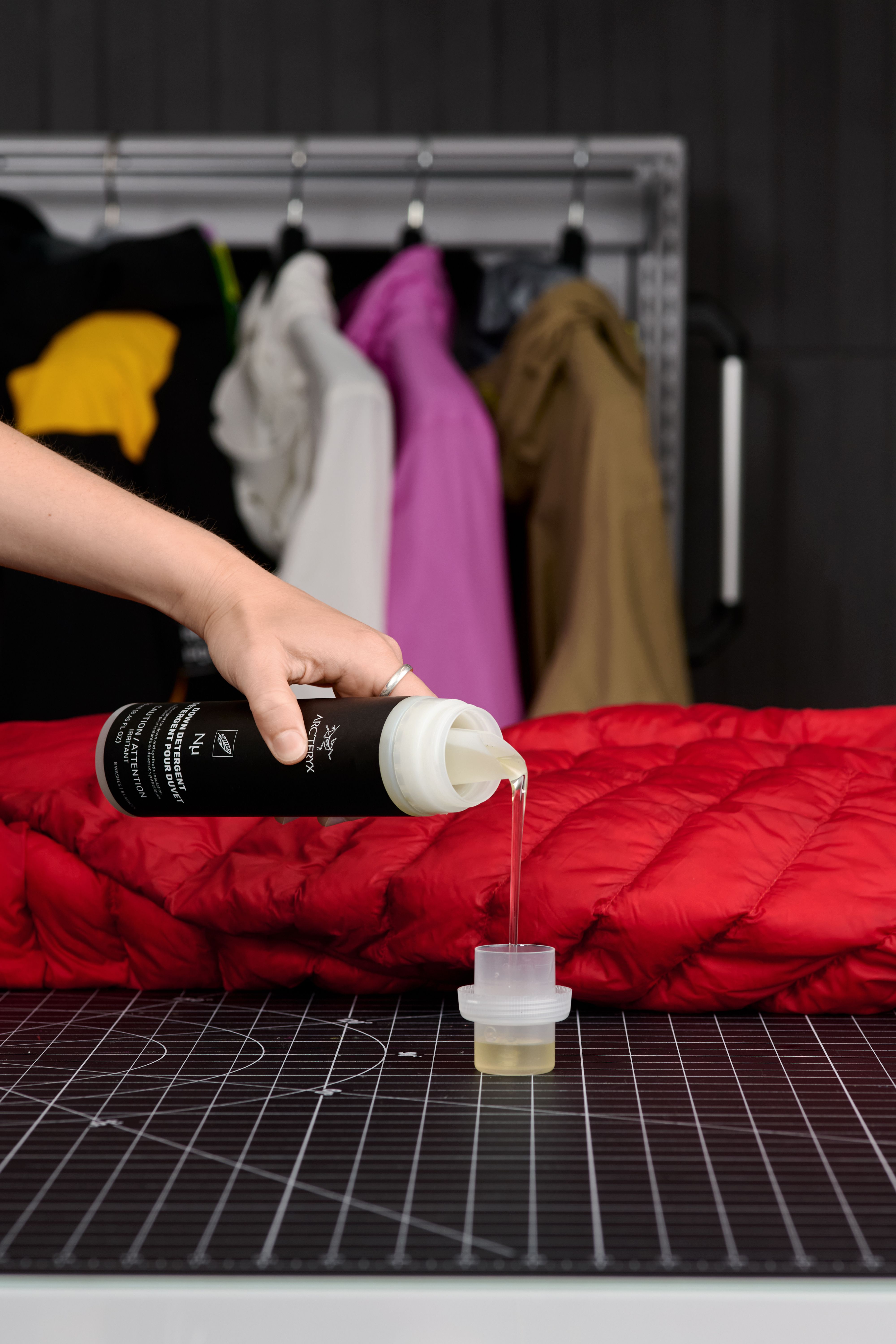 A hand pours Arc'teryx technical wash into a measuring cup beside a red puffer jacket, with other jackets in the background.