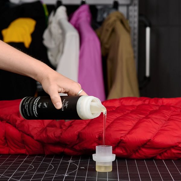 A hand pours Arc'teryx technical wash into a measuring cup beside a red puffer jacket, with other jackets in the background.