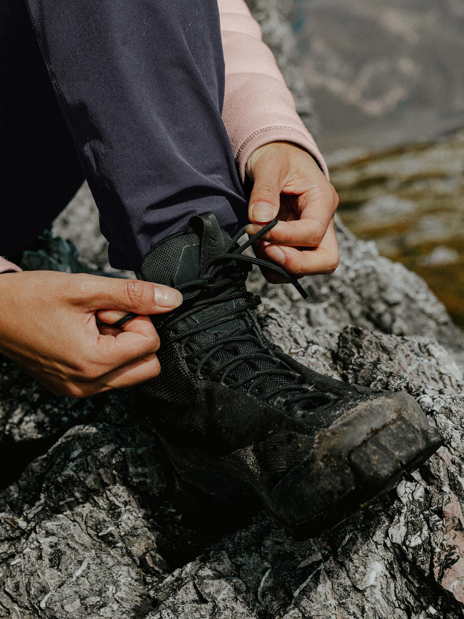 Close-up of a person tying the laces of a black hiking boot on rocky terrain.