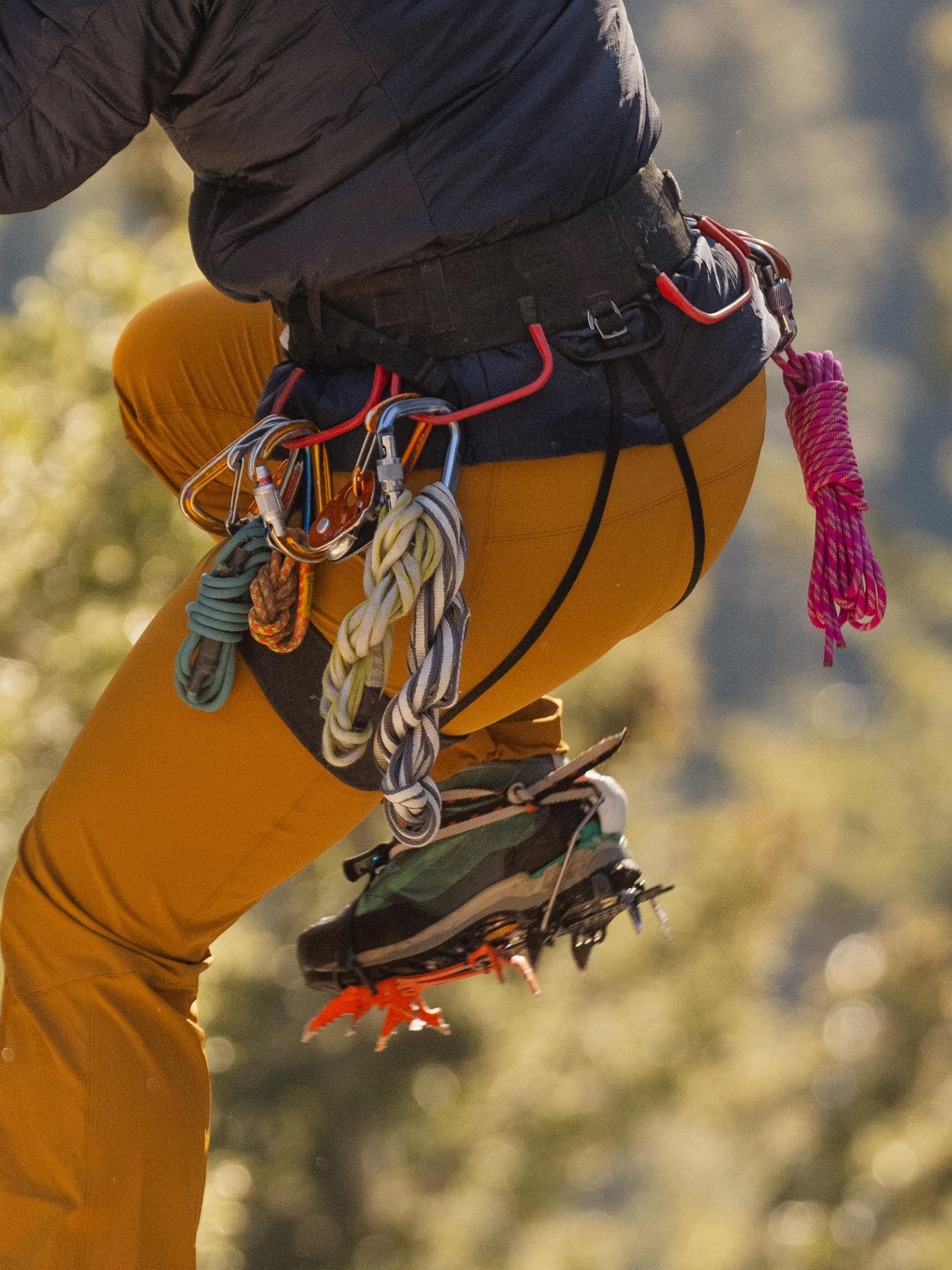 A climber's lower body wearing a harness with carabiners, ropes, and a boot with crampons.