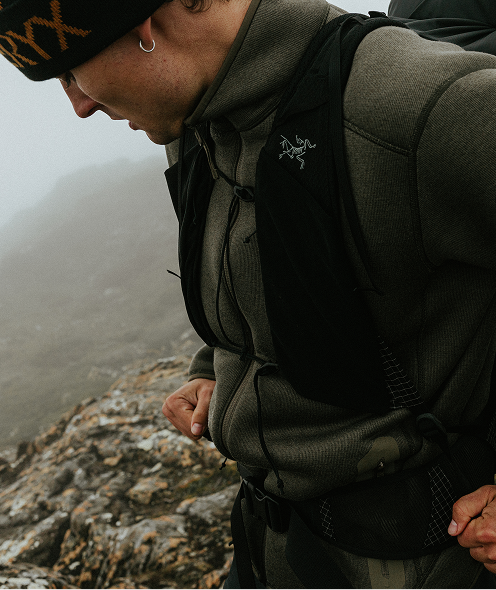 Man in Arc'teryx gear, including a beanie and pack vest, looking down a rocky, foggy mountain.