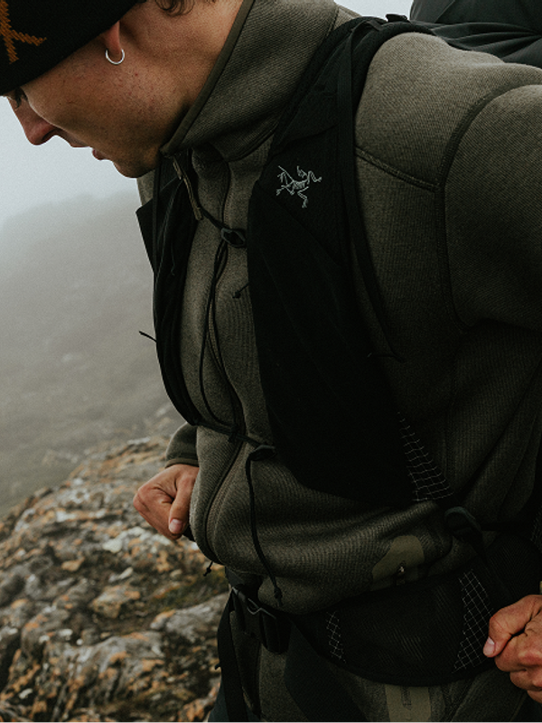 Man in Arc'teryx gear, including a beanie and pack vest, looking down a rocky, foggy mountain.