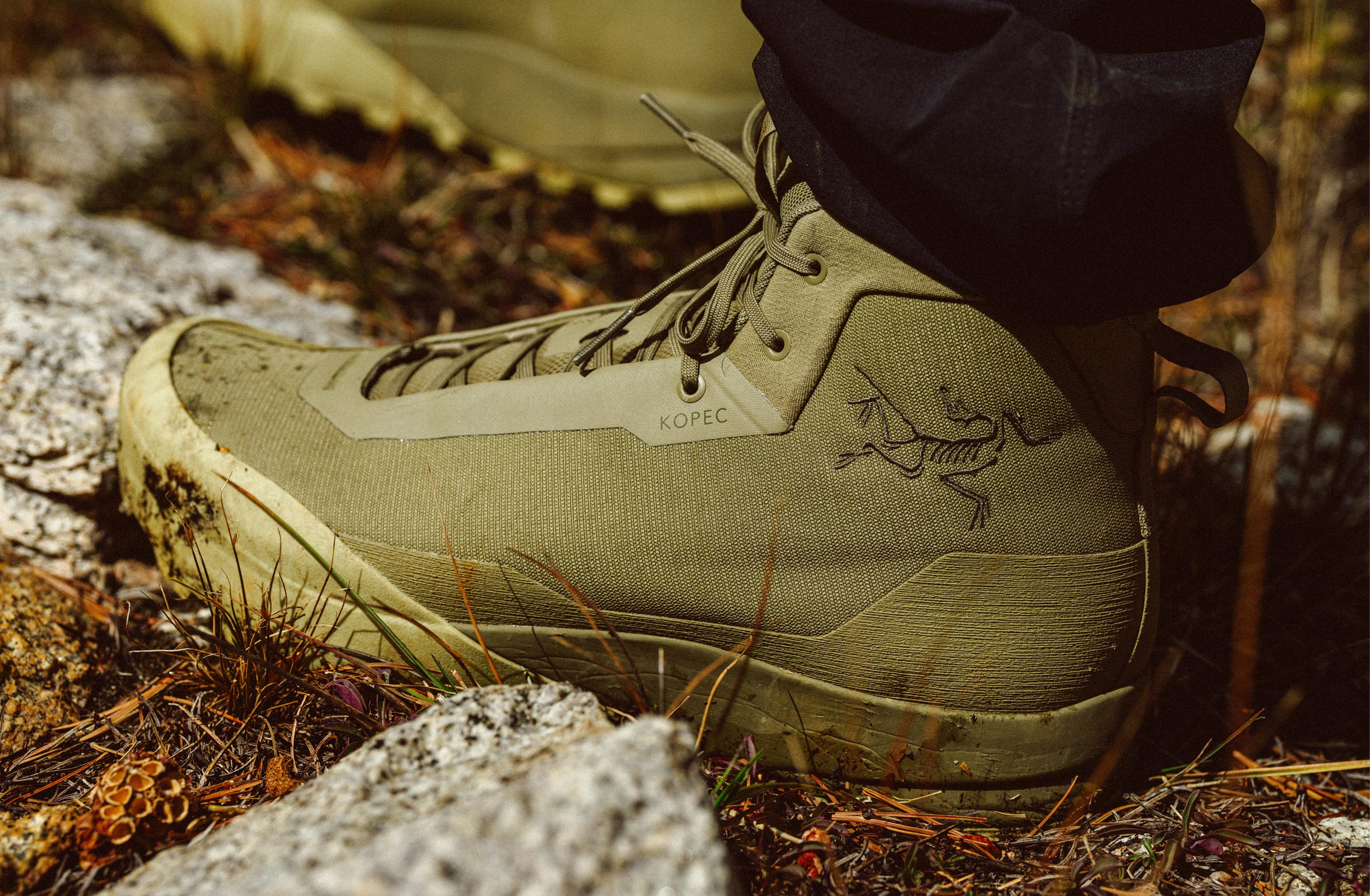 Close-up of a dirty olive-green Arc'teryx hiking boot with "KOPEC" on a tag, worn on rocky terrain.