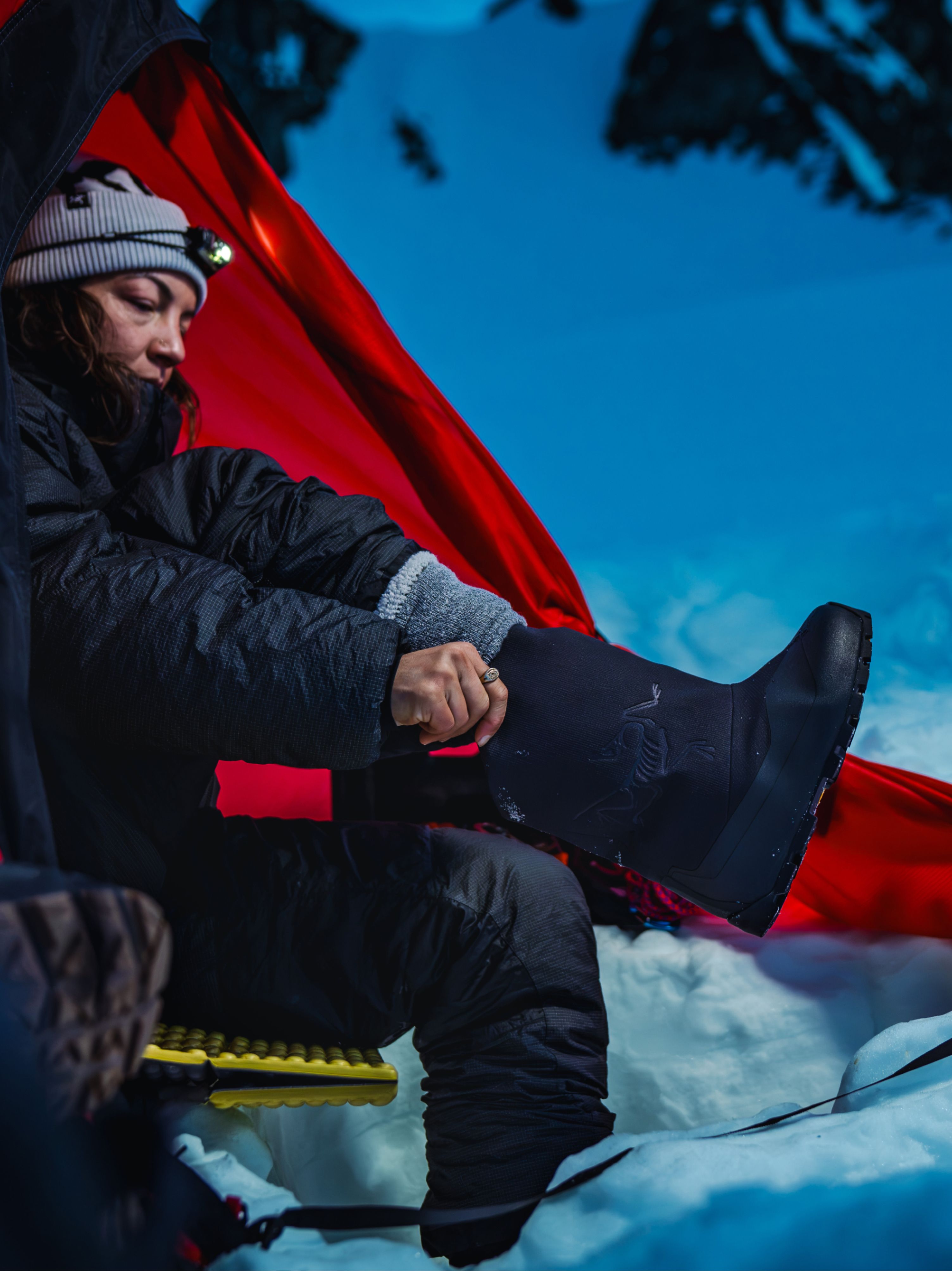 A person in a beanie and puffy jacket puts on a tall black boot while sitting in snow with a red tarp behind them.
