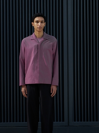 A man in a purple long-sleeve collared shirt and black pants stands against a dark, slatted background.