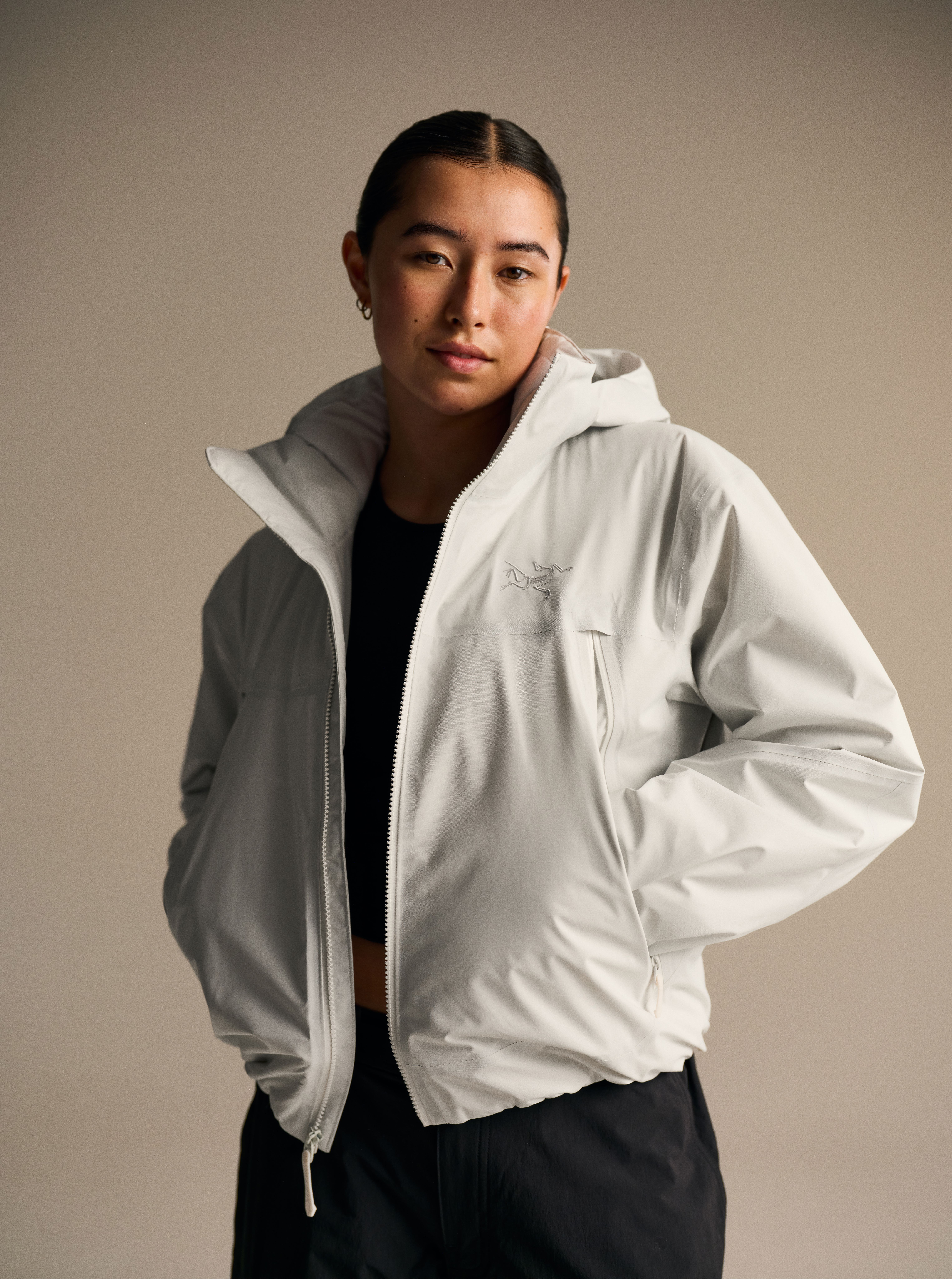 A young woman in a light grey Arc'teryx hooded jacket with hands in her pockets looks at the camera.