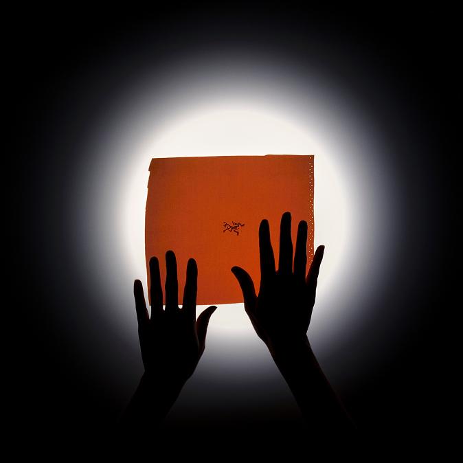 Silhouetted hands hold an orange rectangle with the Arc'teryx logo against a bright circular light on a dark background.