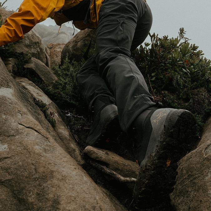 A person in a yellow jacket and dark pants climbs a rocky, vegetated trail, their hiking boots clearly visible.